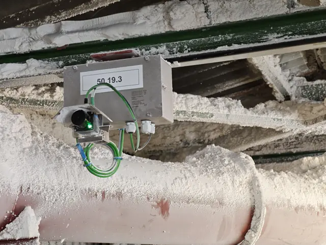 Carcasa de acero inoxidable OWL EYE® VOLUME FLOW con LiDAR 2D y capota protectora, montada en una viga de acero en una planta polvorienta
